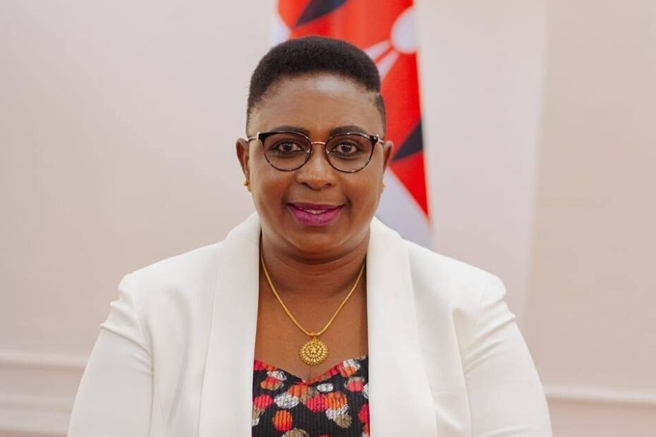 Official portrait of Kenyan politician Aisha Jumwa wearing glasses, a white blazer over a black and patterned top, and a gold pendant necklace, positioned in front of the Kenyan flag.
