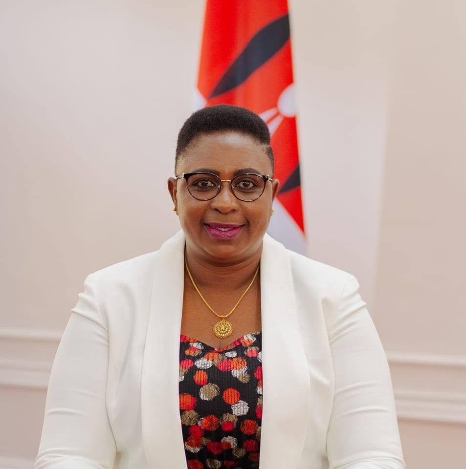 Official portrait of Kenyan politician Aisha Jumwa wearing glasses, a white blazer over a black and patterned top, and a gold pendant necklace, positioned in front of the Kenyan flag.