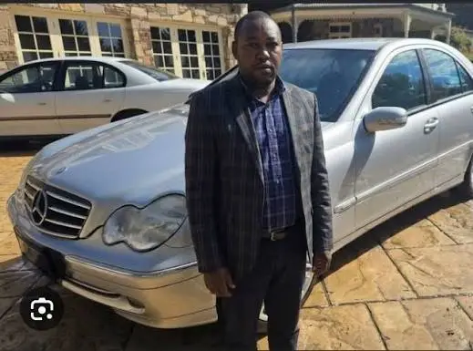 Paul Ngugi Kariuki stands in front of a silver Mercedes-Benz parked on a paved driveway in front of a building.