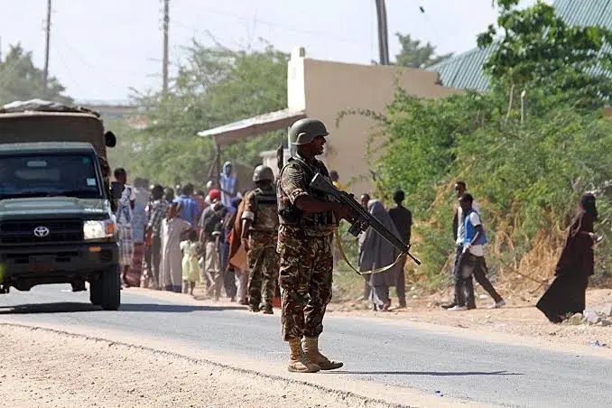 An armed Kenyan security officer standing guard on a paved road next to military vehicles and local residents in Garissa County.
