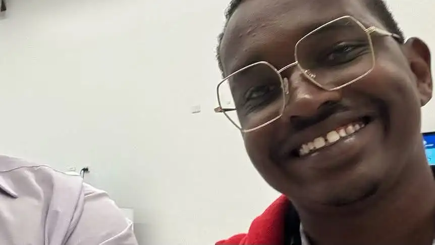Close-up selfie of Alan Kiplimo smiling and wearing wire-rimmed glasses and a red jacket against a plain wall background.