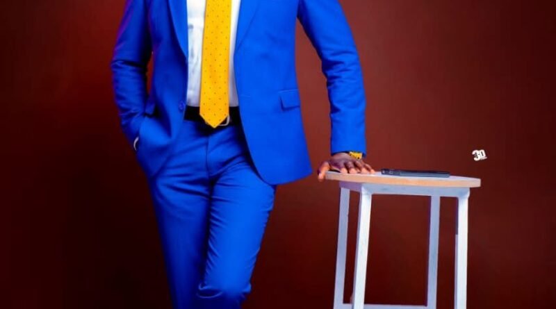 Studio portrait of John Wanyama wearing a royal blue suit and yellow tie while leaning on a white stool.