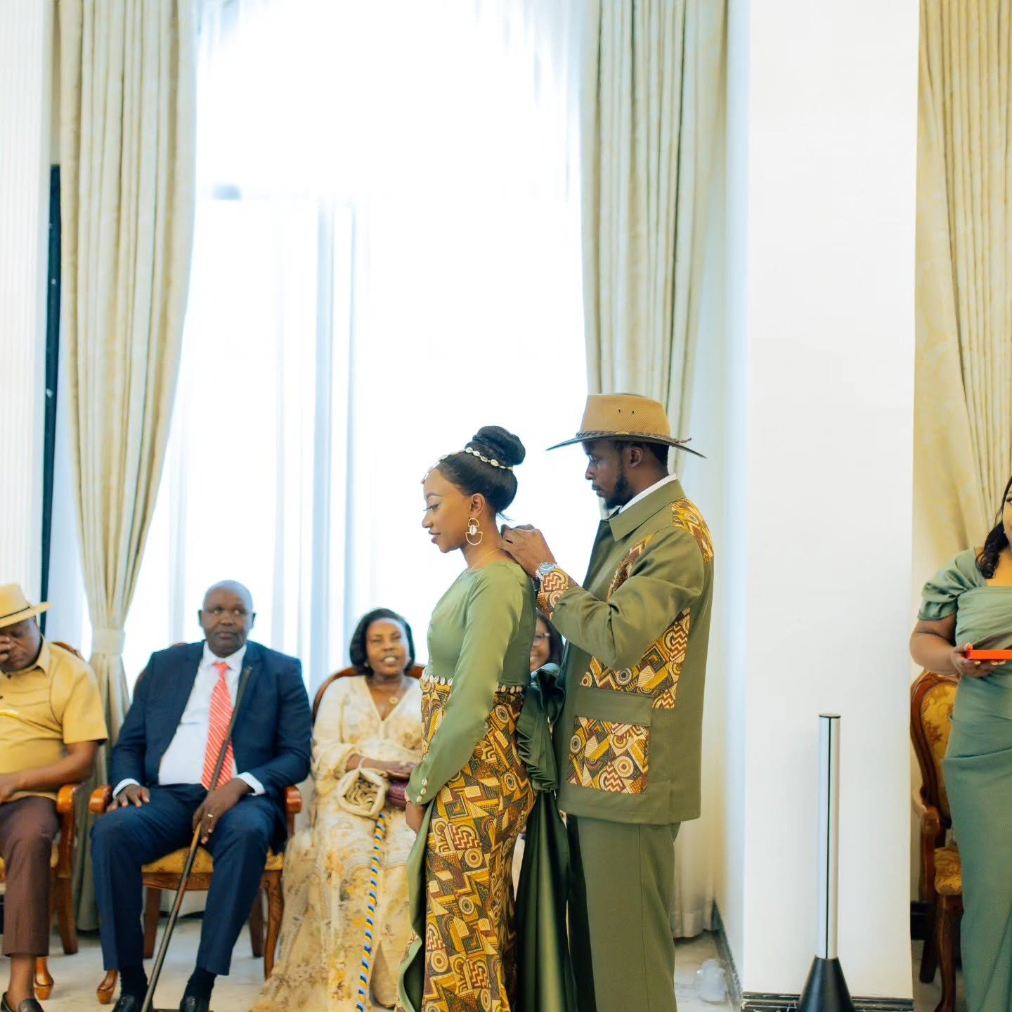 A man in a brimmed hat and olive-green patterned outfit attaches a necklace to a woman dressed in a matching olive-green traditional dress while guests watch seated in the background.