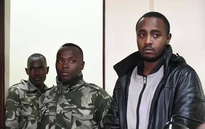 A suspect in a black leather jacket stands inside a courtroom dock flanked by officers in camouflage uniform.