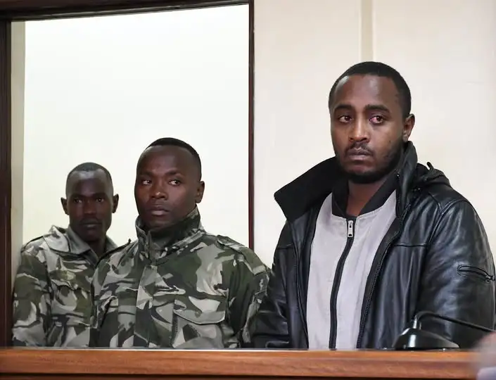 A suspect in a black leather jacket stands inside a courtroom dock flanked by officers in camouflage uniform.
