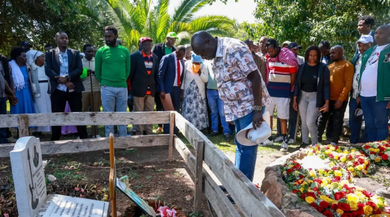 Rigathi Gachagua holding a white hat and bowing his head in respect at a flower-covered grave alongside community mourners in Narok County.