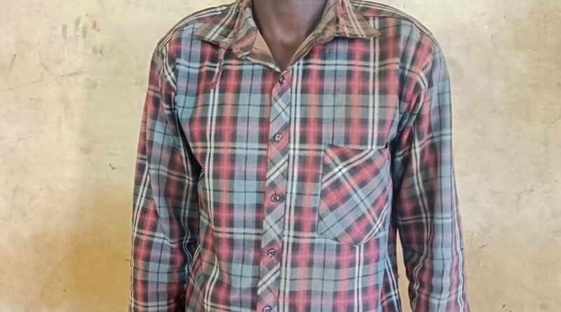 Full-length police mugshot of a young man standing against a beige wall, wearing a long-sleeved red, grey, and black plaid button-down shirt.