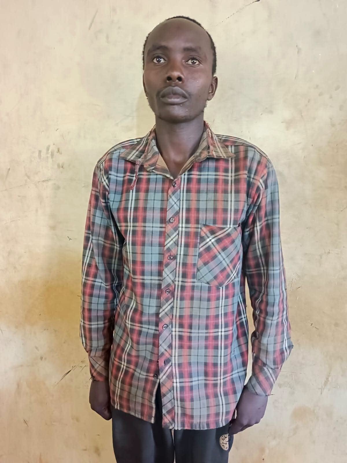 Full-length police mugshot of a young man standing against a beige wall, wearing a long-sleeved red, grey, and black plaid button-down shirt.