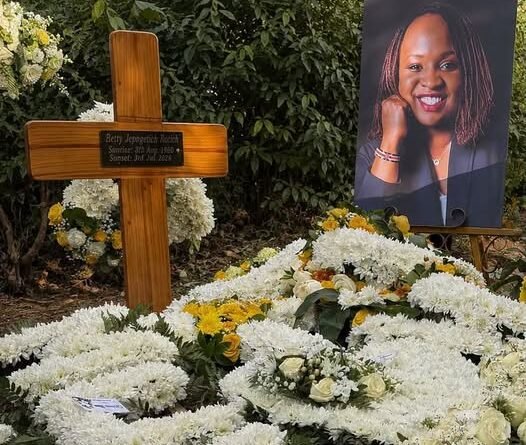 A grave covered in white and yellow floral wreaths, featuring a wooden cross and a portrait portrait on an easel.