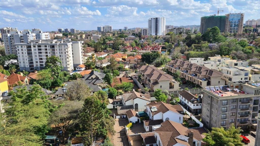 High-angle view of the Kileleshwa neighborhood in Nairobi, featuring modern apartment complexes, townhomes, single-family houses with brown tiled roofs, and lush greenery under a partly cloudy blue sky.