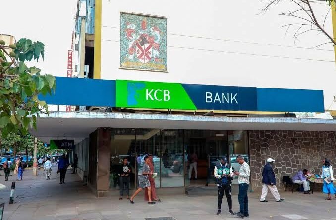 Exterior view of a KCB Bank branch with pedestrians walking on the pavement outside.