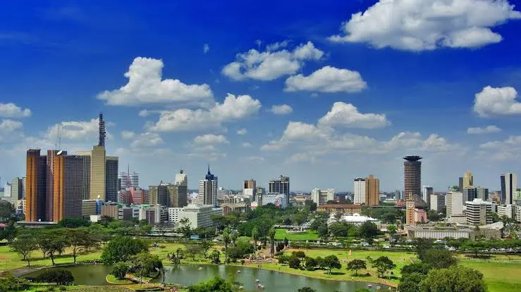 Panoramic view of the Nairobi city skyline and lush green parkland under a bright blue sky with fluffy white clouds.