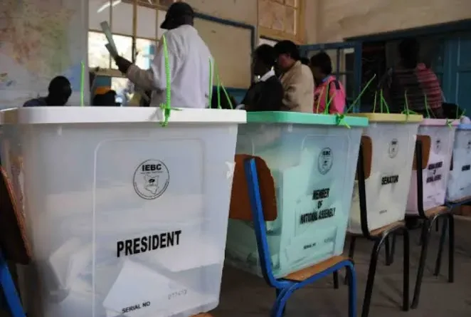 A close-up view of semi-transparent plastic ballot boxes lined up on wooden school chairs inside a Kenyan polling center. The prominent box in the foreground displays the official IEBC seal and is labeled "PRESIDENT" in bold black text. Adjacent boxes are marked "MEMBER OF NATIONAL ASSEMBLY" and "SENATOR". In the softly blurred background, election clerks and officials work near tall classroom windows.