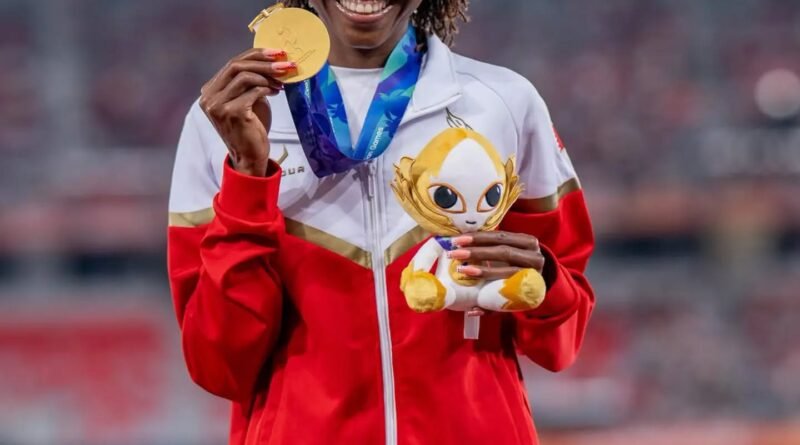 Winfred Yavi smiling brightly while holding up her gold medal and event mascot on the victory podium